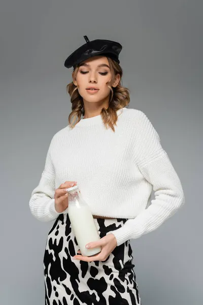 Curly woman in black beret and skirt with cow print holding bottle with milk isolated on grey — Stock Photo