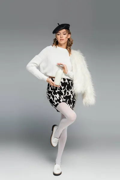 Young woman in beret, faux fur jacket and skirt with animal print holding bottle with milk on grey — Stock Photo