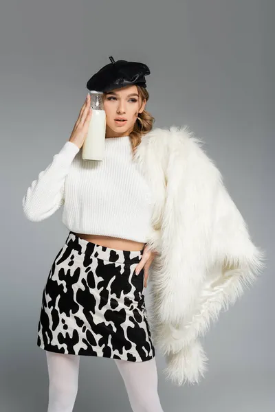 Young woman in beret, white faux fur jacket and skirt with cow print holding bottle with milk isolated on grey — Stock Photo