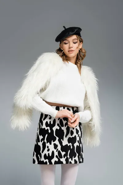 Young woman in beret, white faux fur jacket and skirt with animal print posing isolated on grey — Stock Photo
