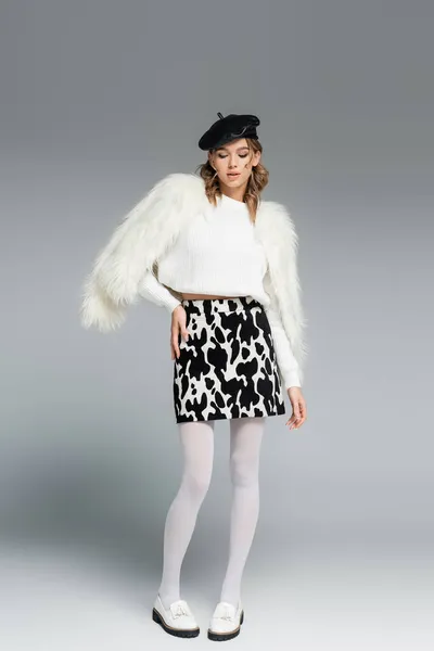 Full length of young woman in beret, white faux fur jacket and skirt with animal print posing on grey — Stock Photo