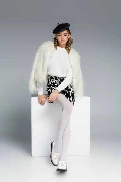 Full length of young woman in beret, white faux fur jacket and skirt with animal print sitting on cube on grey — Stock Photo