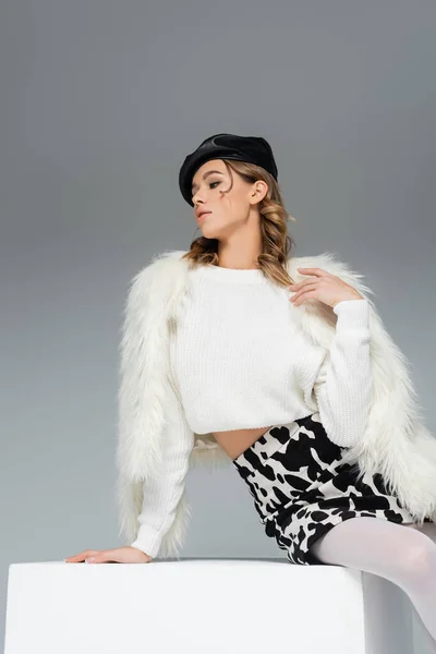 Young woman in beret, faux fur jacket and skirt with animal print posing on white cube isolated on grey — Stock Photo