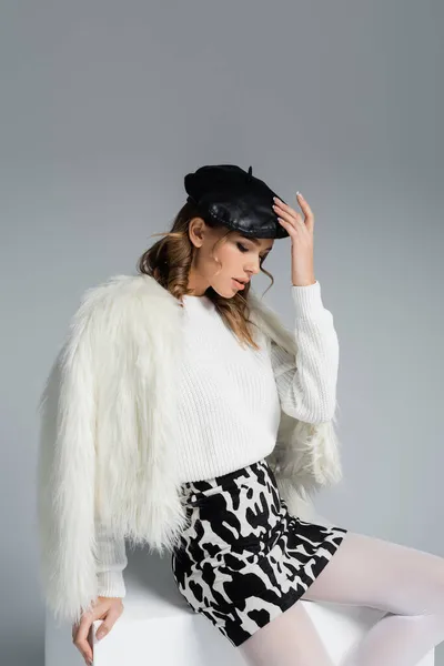 Giovane donna in berretto, giacca bianca in eco pelliccia e gonna con stampa animalier in posa sul cubo isolato su grigio — Foto stock