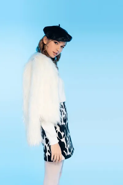 Young woman in beret, white faux fur jacket and skirt with cow print looking at camera on blue — Stock Photo