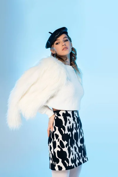 Young woman in black beret, white faux fur jacket and skirt with cow print posing on blue — Stock Photo