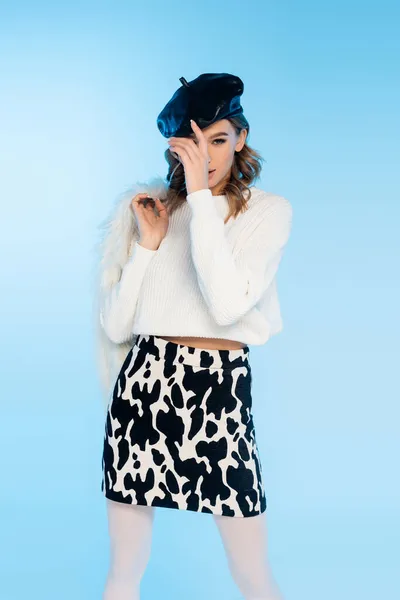 Jeune femme en jupe avec imprimé vache tenant veste blanche en fausse fourrure tout en ajustant le béret sur bleu — Photo de stock