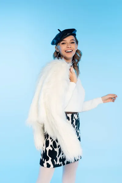 Joyful young woman in beret and skirt with cow print holding white faux fur jacket while posing on blue — Stock Photo