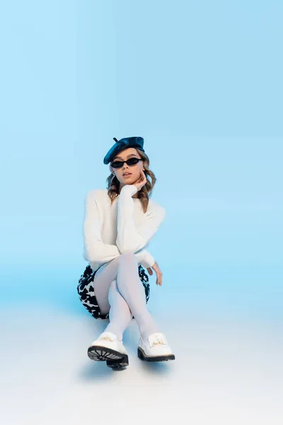 Jeune femme tendance en béret, lunettes de soleil et jupe avec imprimé vache posant sur bleu — Photo de stock