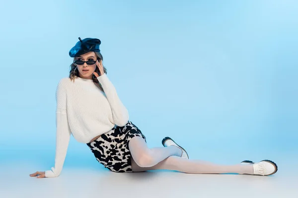 Elegante giovane donna in berretto, occhiali da sole e gonna con stampa mucca in posa su blu — Foto stock