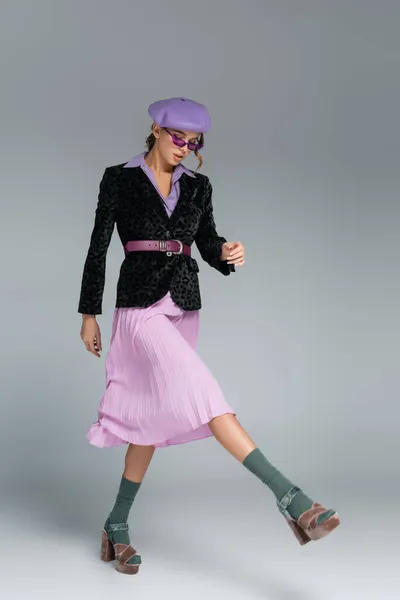 Full length of stylish young woman in beret, purple skirt and blazer with animal print walking on grey — Stock Photo