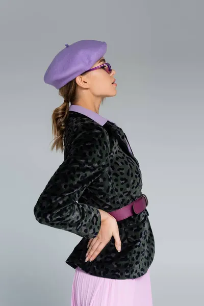 Side view of stylish woman in beret and blazer with animal print posing with hand on hip isolated on grey — Stock Photo