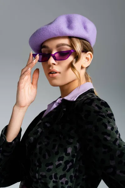 Jeune femme tendance en béret et blazer avec imprimé animal ajustant les lunettes de soleil isolées sur gris — Photo de stock