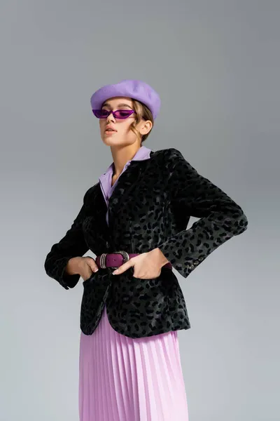 Giovane donna alla moda in berretto, occhiali da sole e blazer con stampa animalier in posa con le mani in tasche isolate sul grigio — Foto stock