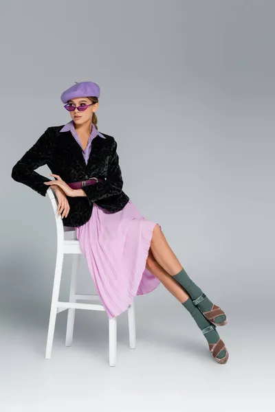Full length of trendy young woman in beret, sunglasses and blazer with animal print posing while sitting on chair on grey — Stock Photo