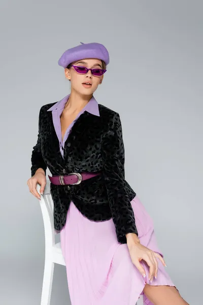 Jeune femme à la mode en béret, lunettes de soleil et blazer avec imprimé animal posant assis sur une chaise isolée sur gris — Photo de stock