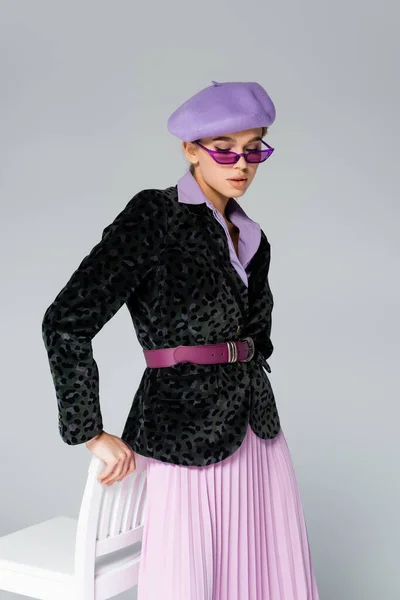 Модная женщина в фиолетовом берете, солнцезащитные очки и блейзер с отпечатком животного позирует возле стула изолированы на сером — стоковое фото