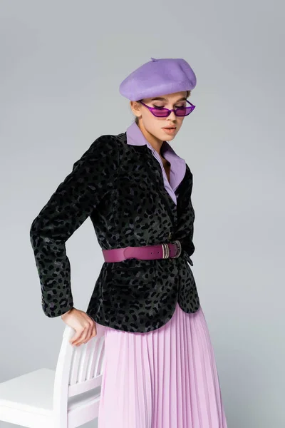 Trendy woman in purple beret, sunglasses and blazer with animal print posing near chair isolated on grey — Stock Photo