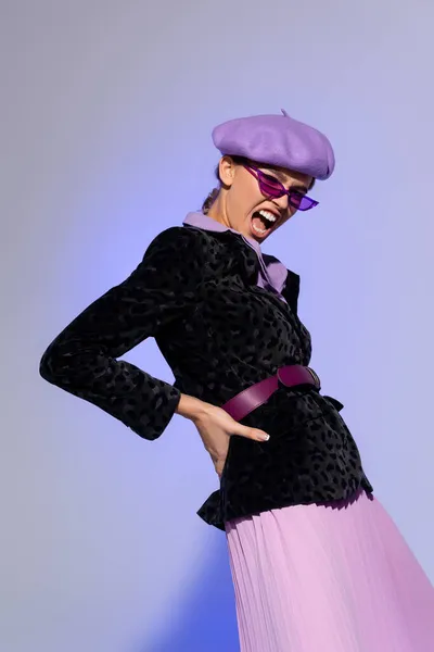 Trendfrau in Baskenmütze, Sonnenbrille und Blazer mit Animal Print posiert mit der Hand an der Hüfte, während sie auf Violett schreit — Stockfoto