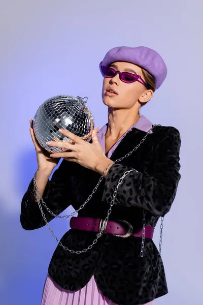 Модная женщина в берете, солнцезащитные очки и пиджак с животным принтом проведение диско мяч на фиолетовый — стоковое фото