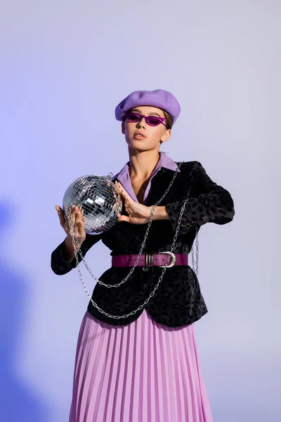 Femme élégante en béret, lunettes de soleil et blazer avec imprimé animal tenant boule disco sur violet — Photo de stock