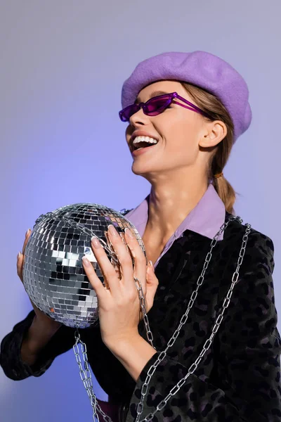 Модная женщина в берете, солнцезащитные очки и пиджак с животным принтом держа диско мяч и смеясь изолированы на фиолетовый — стоковое фото