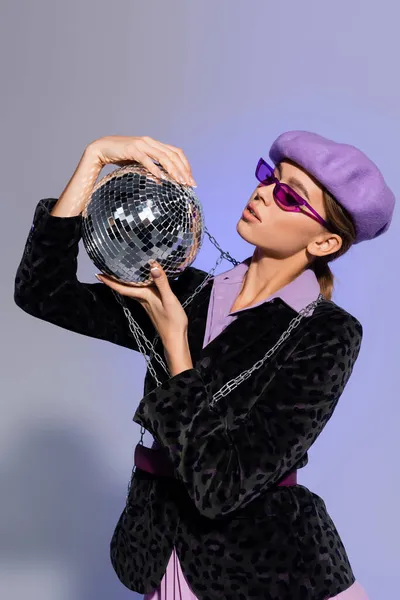 Femme élégante en béret, lunettes de soleil et blazer avec imprimé animal regardant boule disco sur violet — Photo de stock