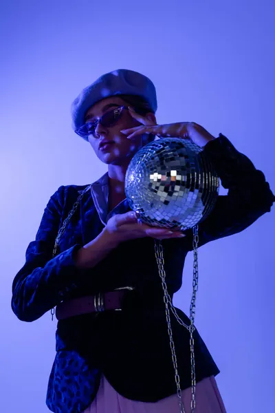 Young stylish woman in beret, sunglasses and blazer with animal print holding disco ball isolated on blue — Stock Photo