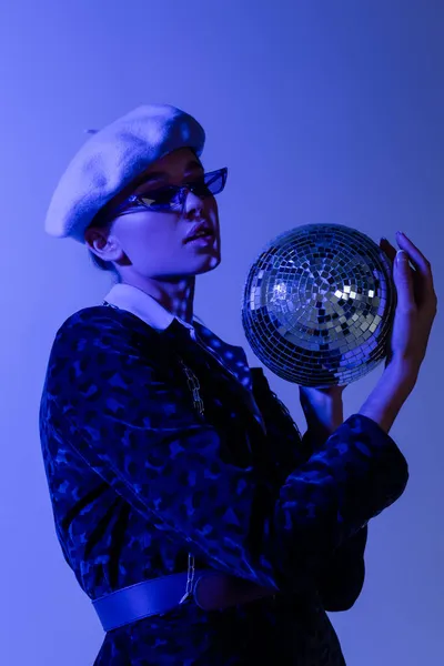 Young woman in beret, sunglasses and blazer with animal print holding disco ball isolated on blue — Stock Photo