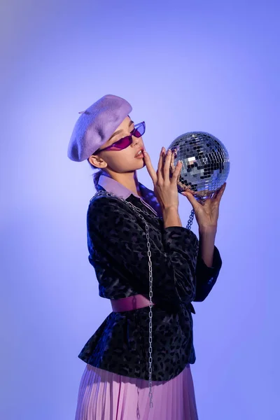 Jeune femme en béret violet, lunettes de soleil et blazer avec imprimé animal tenant boule disco sur violet — Photo de stock