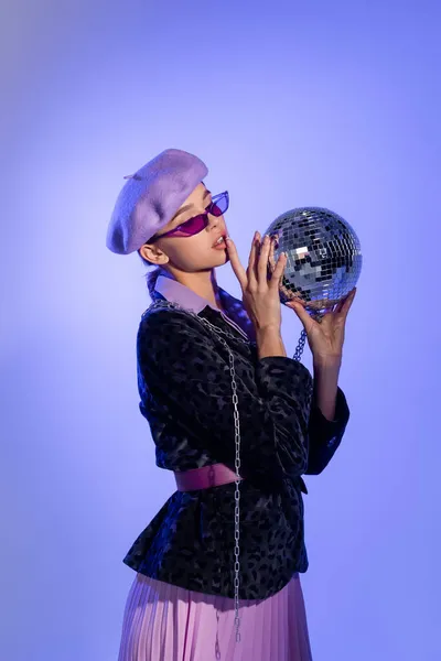 Jeune femme en béret violet, lunettes de soleil et blazer avec imprimé animal tenant boule disco sur violet — Photo de stock