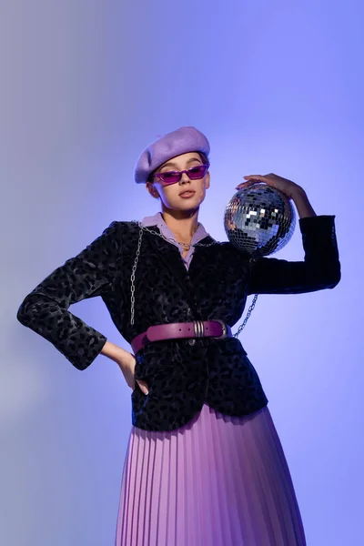 Junge Frau in Baskenmütze und Blazer mit Animal Print hält Discokugel und posiert mit der Hand an der Hüfte auf blau und lila — Stockfoto