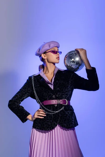 Femme élégante en béret et blazer avec imprimé animal tenant boule disco et posant avec la main sur la hanche sur bleu et violet — Photo de stock