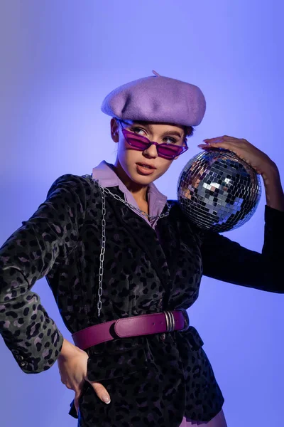 Femme élégante dans des lunettes de soleil et blazer avec imprimé animal tenant boule disco et posant avec la main sur la hanche sur bleu et violet — Photo de stock