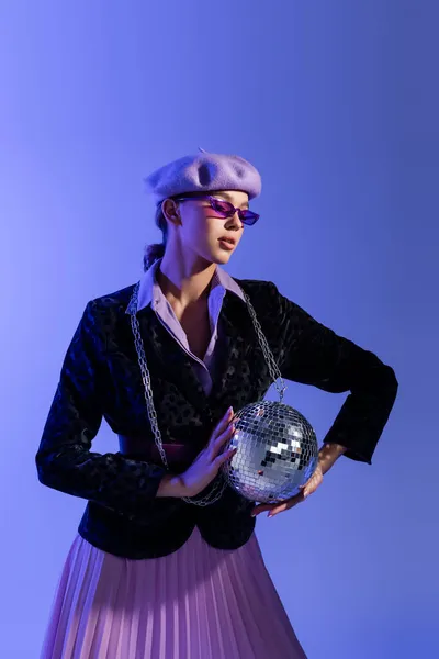 Young stylish woman in trendy sunglasses and blazer with animal print holding disco ball and posing on violet — Stock Photo