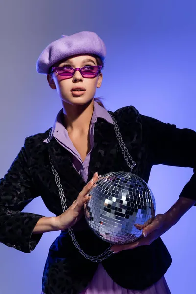 Jeune femme dans des lunettes de soleil à la mode et blazer avec imprimé animal tenant brillant boule disco sur violet — Photo de stock