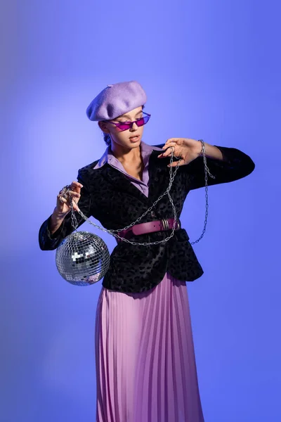Стильна жінка в фіолетовому береті, сонцезахисних окулярах і блістері з тваринним принтом, що тримає диско-кулю на ланцюжку на фіолетовому — Stock Photo