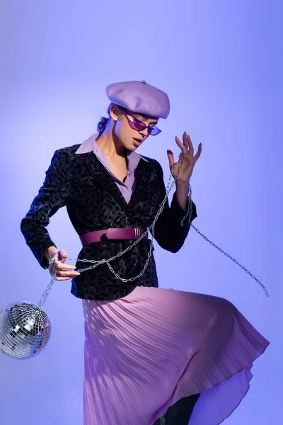 Femme élégante en lunettes de soleil et blazer avec imprimé animal tenant boule disco sur chaîne sur violet — Photo de stock