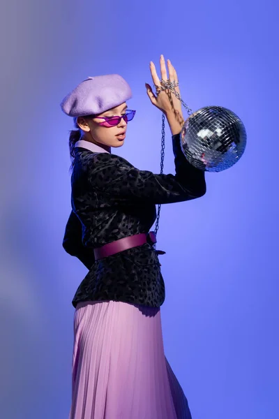 Stylish woman in purple beret and blazer with animal print holding disco ball on chain on violet — Stock Photo