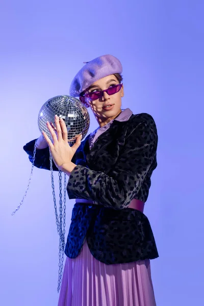 Femme tendance en béret, lunettes de soleil et blazer avec imprimé animal tenant brillant boule disco sur chaîne sur violet — Photo de stock