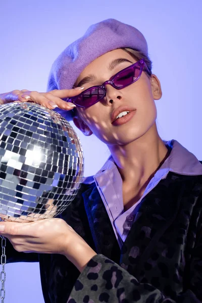 Trendy woman in beret and sunglasses holding disco ball on chain on violet — Stock Photo