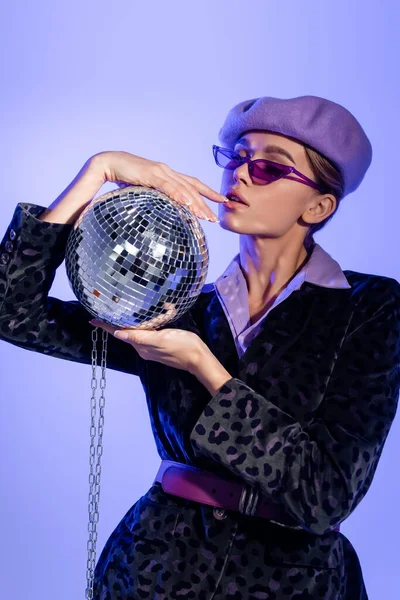 Giovane donna in berretto viola, occhiali da sole e blazer con stampa animalier che tiene palla da discoteca a catena su viola — Foto stock