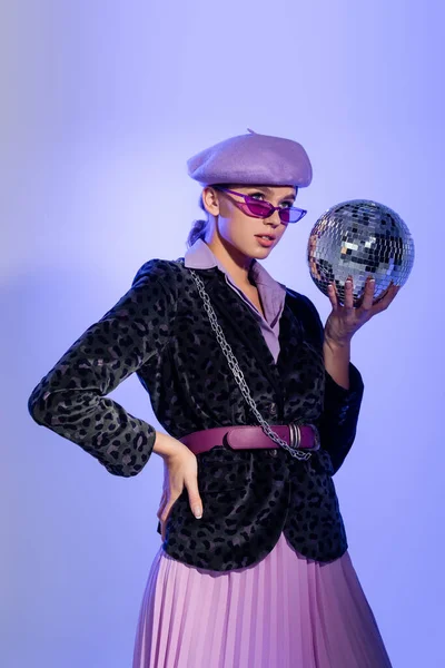 Jeune femme en béret violet et blazer avec imprimé animal tenant boule disco tout en posant avec la main sur la hanche sur violet — Photo de stock