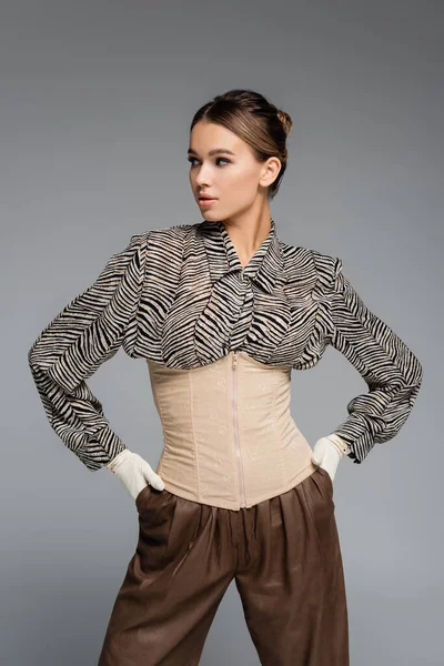 Trendy woman in blouse with animal print, gloves and corset posing with hands in pockets isolated on grey — Stock Photo