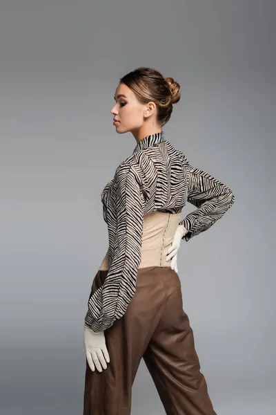 Femme à la mode en chemisier imprimé animal, gants et pantalon marron posant à la main sur la hanche isolé sur gris — Photo de stock