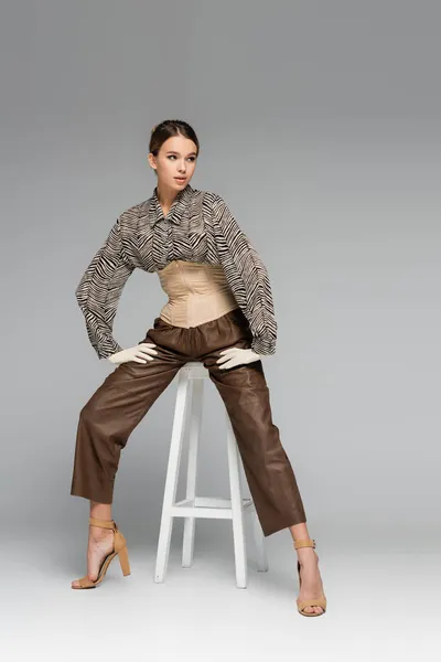 Full length of trendy young woman in blouse with zebra print sitting on stool on grey — Stock Photo