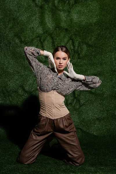 Stylish woman in blouse with zebra print, corset and gloves standing on knees near grassy background — Stock Photo