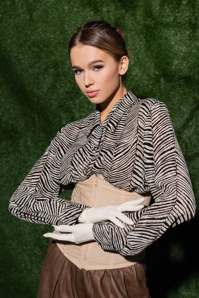 Modelo de moda joven en blusa con estampado de cebra, corsé y guantes mirando a la cámara cerca de fondo cubierto de hierba - foto de stock