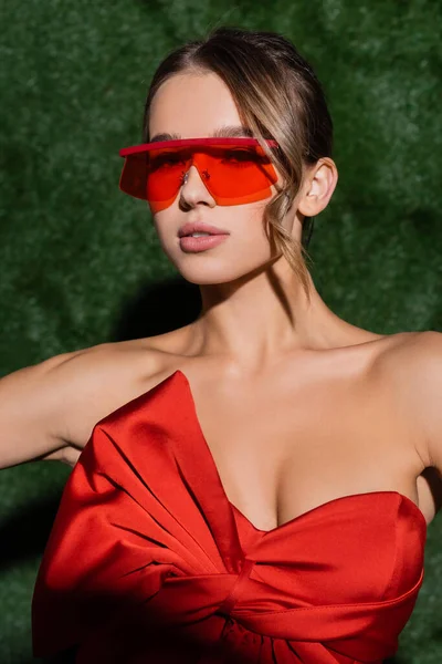 Jeune femme en chemisier rouge et lunettes de soleil regardant la caméra près de fond herbeux — Photo de stock