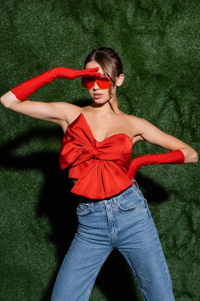 Jeune femme en chemisier rouge, jeans, lunettes de soleil et gants posant à la main sur la hanche près d'un fond herbeux — Photo de stock